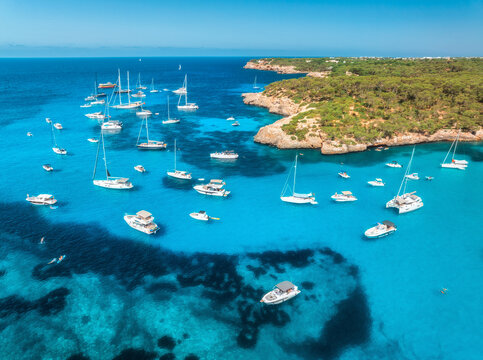 Aerial view of luxury yachts and boats anchored in turquoise sea bay surrounded by rocky coastline and green forest, summer vacation scene in Mallorca, Balearic Islands, Spain. Top drone view of boats