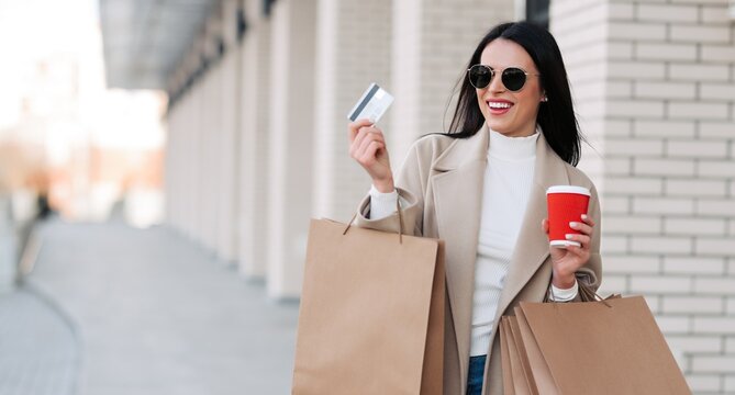 Happy young woman shopping with credit card, coffee and paper bags in a city street