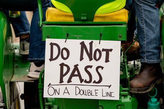 Handmade "Do Not Pass On A Double Line" sign is mounted on a green tractor with a person's boot visible.