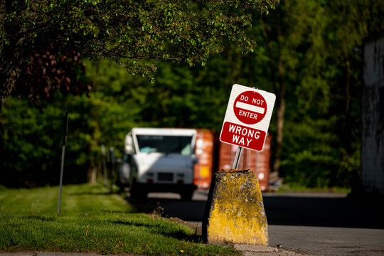 A Do Not Enter sign above a Wrong Way sign stands on a green post at the edge of a suburban road, a US Postal Service mail truck visible but soft in the blurred background.
