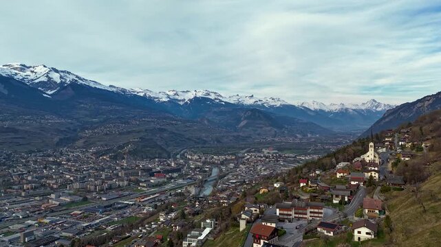 Aerial timelapse over Sion in Rhone Valley, Switzerland, with evening light illuminating alpine town, river and snow capped Swiss Alps. Dramatic scenic mountain landscape. Hyperlapse shot.