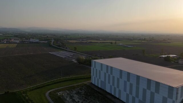Modern data center building in rural Po Valley landscape with adjacent solar farm under construction and distant Apennines forming hazy background under warm sunset light