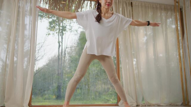 woman exercise indoors in bright room near window, stretching arm and leg on yoga mat with calm posture, standing with open arms and bending forward in wide leg stance