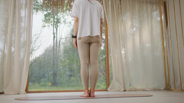 Indoor yoga practice with flexible woman bending forward on mat near window and curtain, standing woman back view in home studio, arms outstretched yoga stretch exercise routine in calm natural light