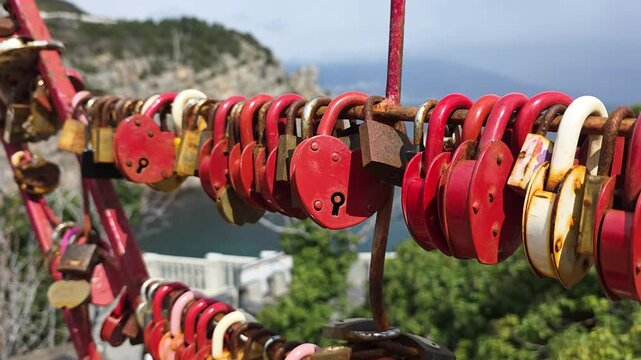 Red and rusted heart-shaped love padlocks symbolize everlasting commitment and romance, securely fastened to a bridge railing with an ocean vista in the background