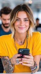 Fototapeta premium Young woman with blonde hair and blue eyes smiles at the camera while holding a black smartphone. She wears a yellow t-shirt, beaded necklaces, and shows arm tattoos in an indoor setting