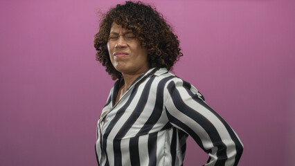 Obraz premium Woman in striped shirt holding lower back with grimacing expression in vibrant pink studio; discomfort.