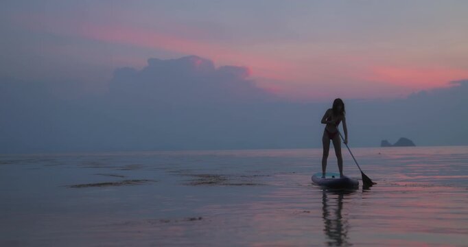 Slim silhouette of trained sportswoman rows SUP in tranquil sea against purple cloudy sky. Lady tourist balances in drifting surfboard in ocean after sunset