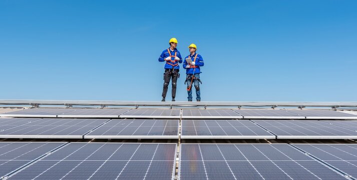Two engineers in hard hats and safety gear inspect solar panels on a rooftop.