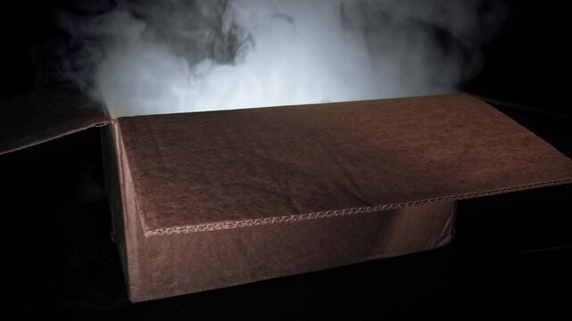 Cardboard box emanating smoke against dark background creating mysterious and intriguing visual effect. Light source hidden inside box illuminates smoke from below, creating atmospheric and dramatic s