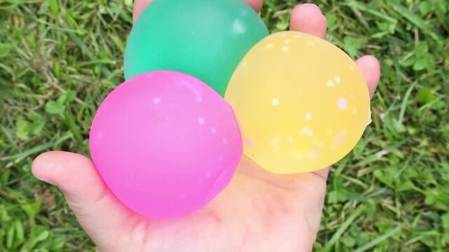 Hand holds and squeezes three colorful squishy antistress toys green pink and yellow together on grass outdoors creating satisfying ASMR tactile experience