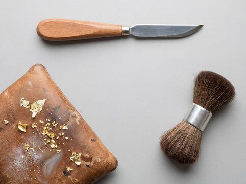 Traditional gilding tools with gold leaf flakes on leather cushion knife and brush flat lay