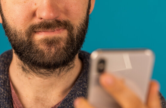 Close up of a bearded man smiling while looking at a smartphone screen, against a vibrant blue backd