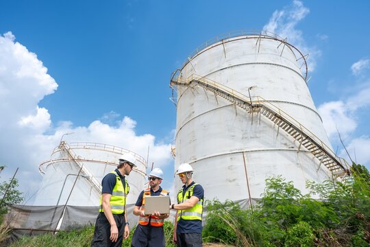 Group of petroleum engineers hold tablet and using radio communication while pointing and discussing project at crude oil storage tanks in refinery site during daytime industrial energy teamwork.