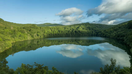 Naklejka premium Breathtaking view of tranquil forest lake surrounded by vibrant green hills with fluffy clouds reflecting on surface of peaceful pond under bright daylight creating perfect natural harmony scene.