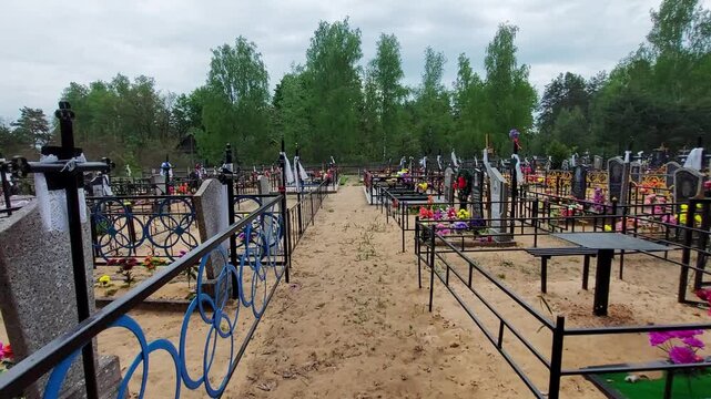 Overview of an Orthodox cemetery showing rows of graves, each enclosed by decorative metal fences. Sandy ground path leads through many headstones, vibrant artificial flowers mark resting places, back