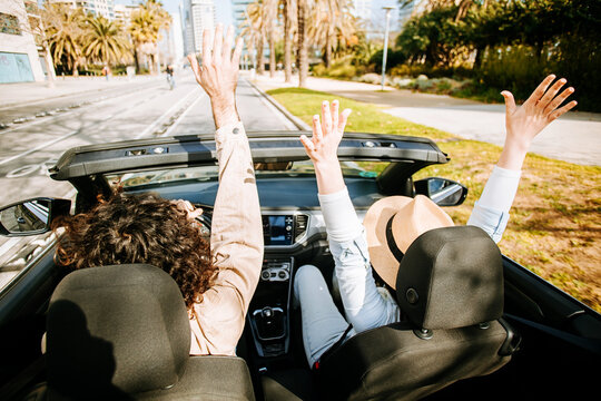 Young adult couple raising hands while riding in convertible car at city street. Man and woman driving while enjoying sunny summer journey together in Barcelona. Travel and lifestyle concept.