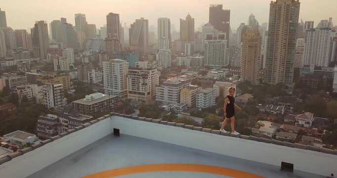 Drone view from helipad rooftop in Bangkok showing sunset skyline warm light over dense high rise buildings wide city panorama and elevated urban perspective with lone person