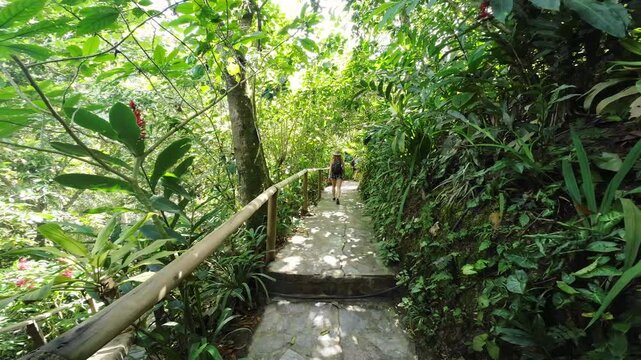 Cinematic shot of a woman with a backpack walking along a lush jungle path in Minca. Golden sunlight through the trees during a peaceful forest bathing walk. Healing nature and wellness.