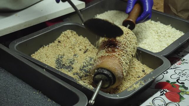 Close up view of person in blue glove rotating chimney cake dough cylinder and applying crushed nuts. Caramel glaze coating, sweet dessert, market environment, traditional snack.