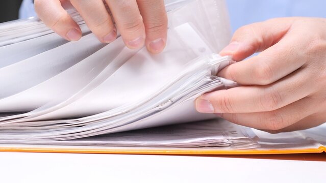 Hands flipping through clear plastic file sleeves in a binder, organizing and searching paperwork to manage documents, records and office files for orderly information storage and administration