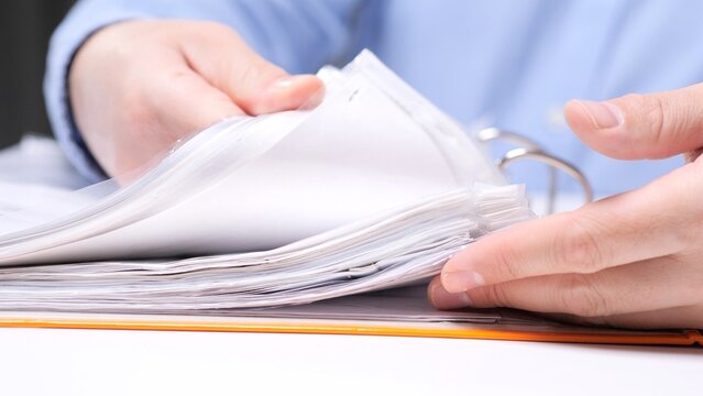Person's hands organizing and reviewing paperwork in a binder, browsing through important documents housed in clear sheet protector sleeves for archiving and office management
