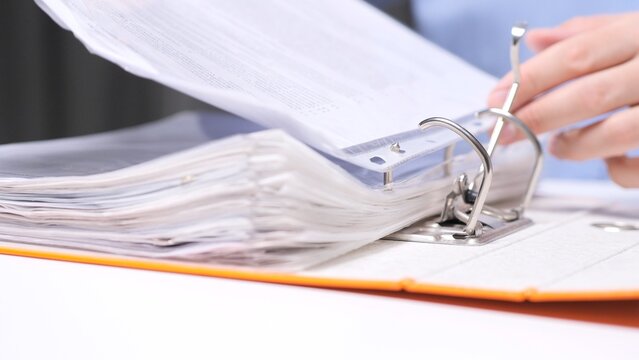 Person's hand inserting document pages into an orange ring binder, organizing paperwork and archiving records at a desk in a professional office setting, close up detail