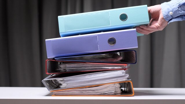 Human hand arranging an extensive pile of multicolored office ring binders filled with paperwork, visualizing concepts of archiving, organization, bureaucracy, and administrative tasks