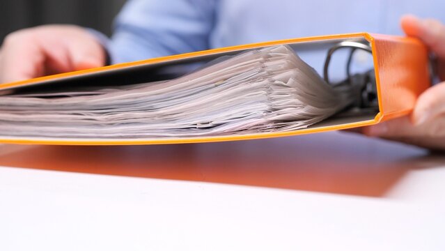 Person holding an overstuffed orange ring binder with overflowing papers and visible folders, symbolizing office paperwork, records management, administrative overload and document organization