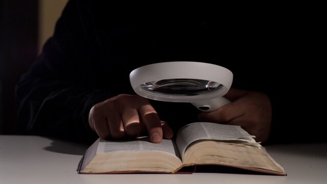 Person holding a magnifying glass over an open old book, carefully examining text and pages in a dark setting, indicating research, education, and investigation