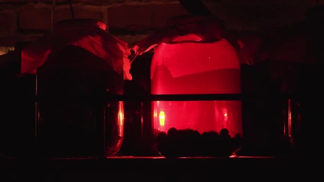 Close up view of two large glass jars in dark room. One jar glows with bright red light, creating ominous atmosphere. Small container of dark, round objects, likely berries, in front of illuminated ja