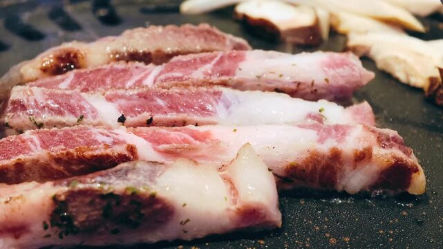 Close-up of thick-cut Korean pork belly (Samgyeopsal) grilling on a black griddle. Slow motion