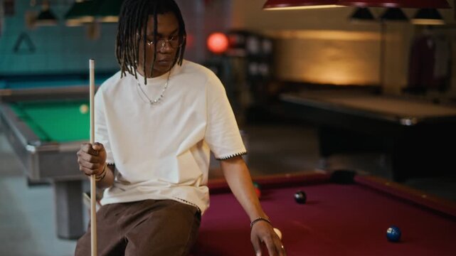 Portrait shot of young adult African American man holding pool cue while leaning on billiard table at snooker hall