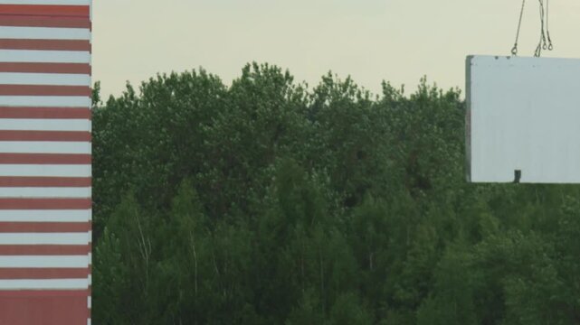 Heavy white block or precast panel suspended by chains of large crane, gently lowered or moved across an outdoor construction site. Dense green trees of forest in background, alongside a distinctive r