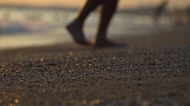 Detailed shot of shimmering sand particles on beach as small ocean wave washes over shore line. Blurred person stands in background enjoying tranquility of nature. Low angle