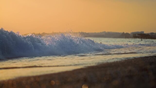 Sea wave breaking on pebble beach at golden hour. Warm light reflecting on water surface creating shimmering effect. Blurred bathers enjoy calm sea conditions. Vacation holiday leave sea. Slow motion.