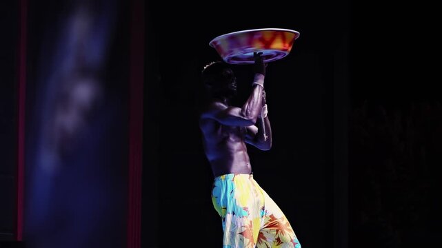 Skillful black African male artist engages audience with captivating balancing act, holding vibrant bowl steady on stick with two hands, shirtless upper body and patterned pants visible against deep s