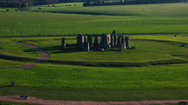 A sweeping aerial drone shot of the iconic Stonehenge prehistoric monument in Wiltshire, England, captured during the golden hour. The ancient standing stones and circular earthworks