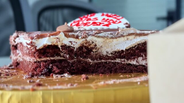 Close-up of a delicious sliced chocolate cake with cream layers and frosting. The dessert sits on a gold base in a bright indoor setting, showcasing rich textures and sweet details.