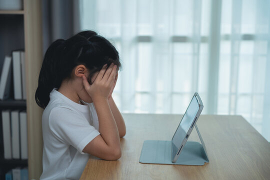Young girl feeling frustration and sadness while looking at a tablet device on a wooden desk in a well-lit room with soft curtains in the background