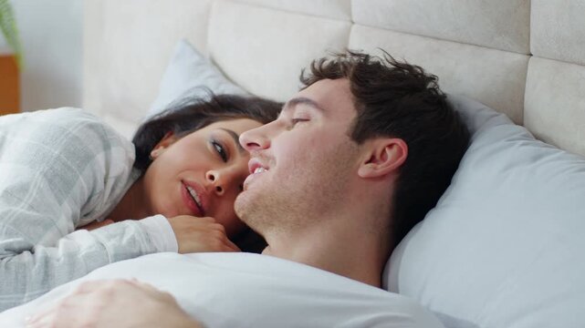 Happy young Caucasian couple lying together on bed bedroom cuddling talking love weekend comfortable man woman girl guy chatting smiling sweetheart tenderness rest relax homeowner relationship tenant