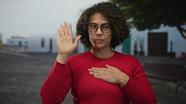 Woman with glasses raises right hand and places left hand on chest in street; commitment sincerity calm.