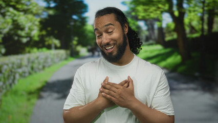 Fototapeta premium Man with hand on chest smiling on forest path wearing white tshirt, bearded with curly ponytail in green park outdoors; gratitude humble.