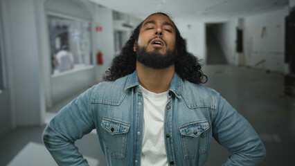 Obraz premium Man with beard and long hair clutching chest and closing eyes in empty studio space, denim jacket visible, hand pressed to sternum, possible heartattack; pain vulnerability struggle recovery.