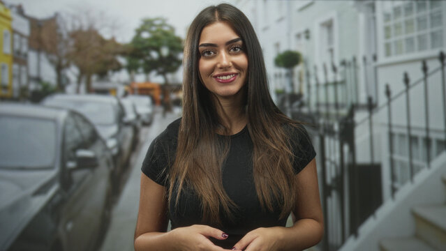 Woman smiling, hands clasped at waist, facing camera on a residential street with parked cars and iron railings; confidence.