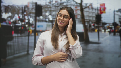Fototapeta premium Woman young hispanic wearing glasses smiling, looking up with hands clasped on a busy rainy city street; joy hope.