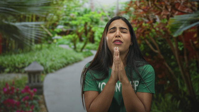 Woman volunteer in green t shirt with hands pressed in prayer gesture and pleading facial expression on a garden path in forest; hope plea.