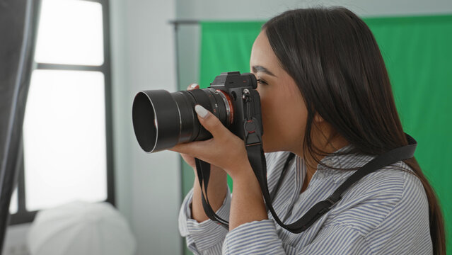 Woman holding camera with hands on lens, looking through viewfinder and later checking playback while smiling in studio with green screen and softbox; creative joy.