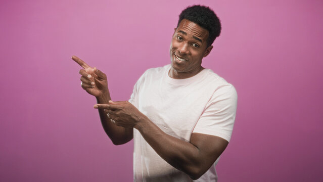 Man pointing with fingers and smiling, bare hands visible wearing a pale t shirt in studio with pink backdrop; playful confidence.