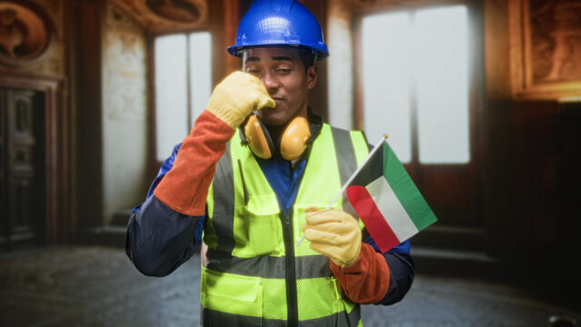 Man construction worker holding kuwaiti flag and raising gloved hand in building interior; pride unity celebration.